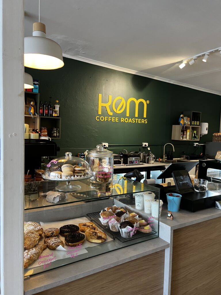 Golden yellow KOM Coffee Roasters logo on the deep green wall behind the counter