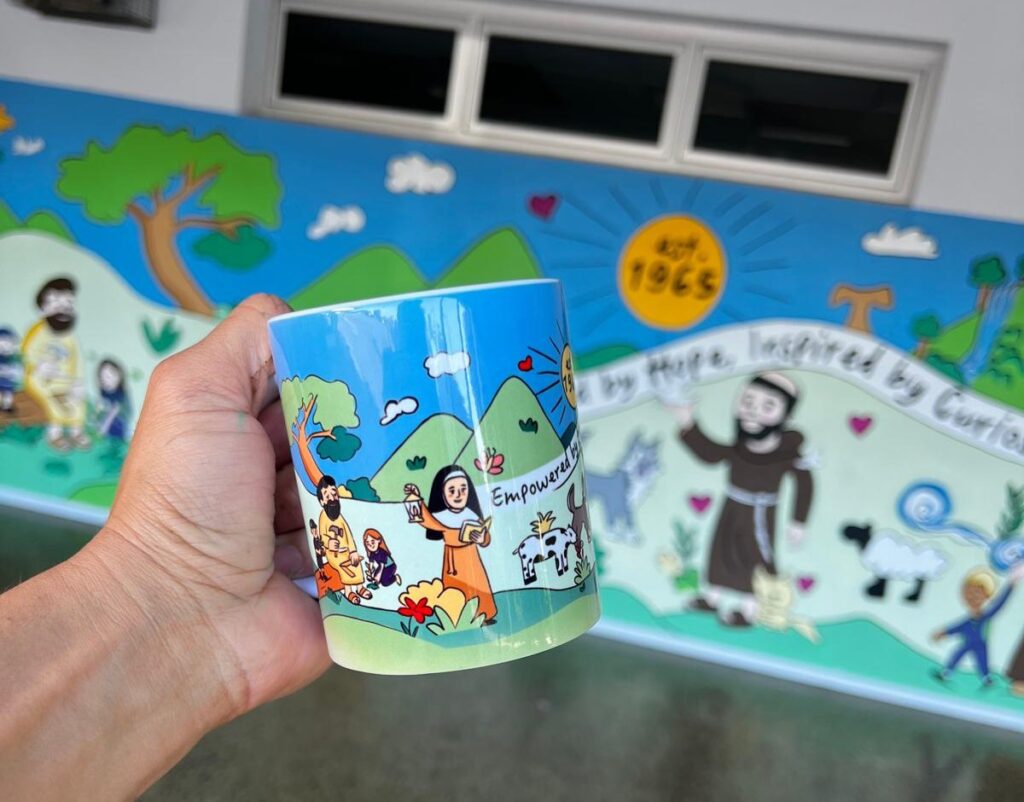 Custom mug printed with the St Francis of Assisi School mural design, held in front of the mural
