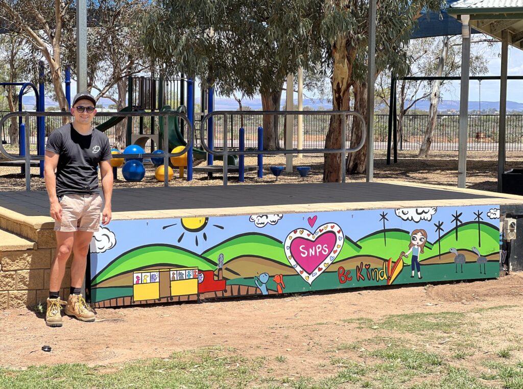 Diegodalo standing next to the finished hand-painted mural panels on the Stirling North Primary School stage
